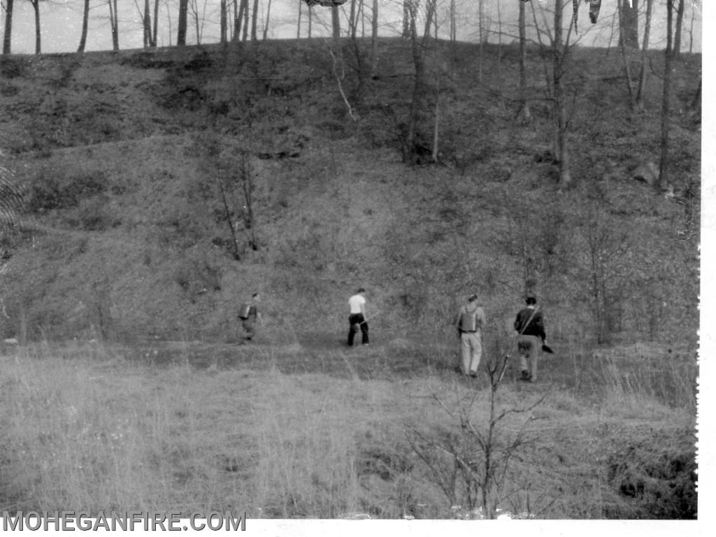 Members walk with brooms and indian tanks to put out a brush fire in the woods 